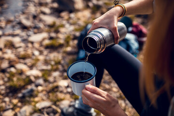 Close up of a young woman pouring warm water from a thermos into a mug filled with coffee while out hiking in nature