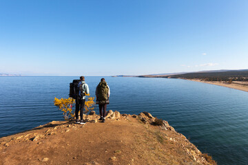Fototapeta na wymiar Baikal Lake in Autumn. Tourists with backpacks travel around Olkhon Island on foot and admire the beautiful view of Saraisky Bay from the cliff. Autumn active outdoors and recreation