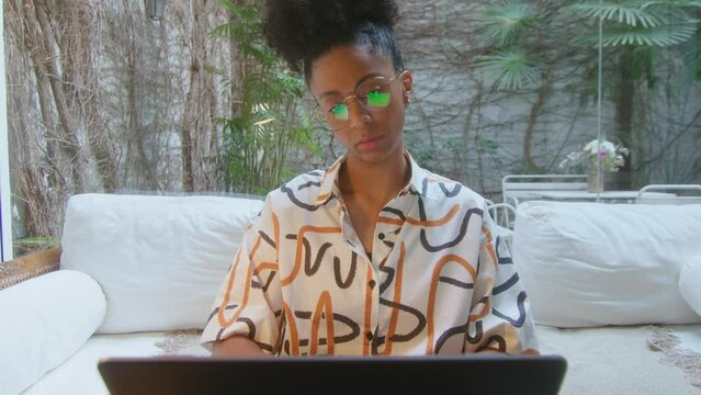 African-American woman sitting in modern home office with backyard garden view, opening laptop and start typing. Crash zoom shot