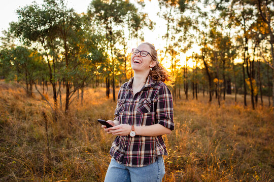 Happy young woman holding phone laughing in rural farm paddock