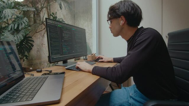 Concentrated programmer in wireless earphones sitting at desk in modern home office and writing code on a computer and laptop. Whip zoom shot