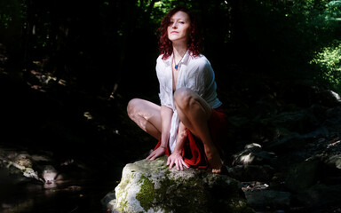 Proud confident adult sexy woman sitting arms between legs spread on stone in creek bed in nature with red dyed hair