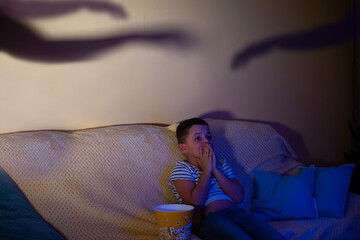 A little boy watches a horror movie while sitting on the sofa with popcorn, covering his mouth with...