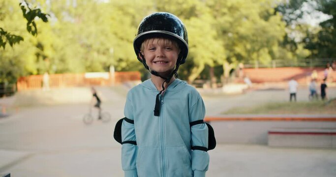 Portrait of a happy blond boy in a blue jacket in a black helmet and elbow pads, who stands against the backdrop of a skatepark in the park