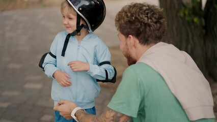 A little blond boy in a blue sweater does not want to wear a black protective helmet for riding a skateboard in a skate park, which his father with curly hair in a Green T-shirt put on him