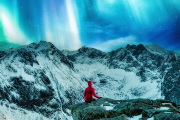 Mountaineer in red jacket sitting on the cliff with Aurora borealis over snowy mountain on Ryten in Lofoten Islands