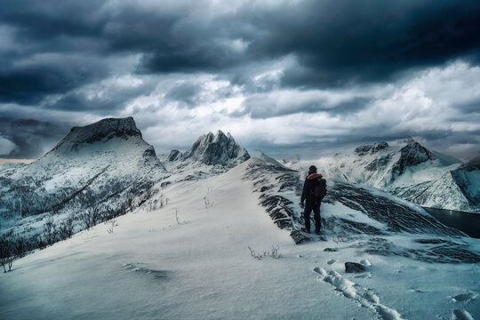 Mountaineer standing on top of snowy mountain with dark stormy sky in winter on Segla mount at Senja Island