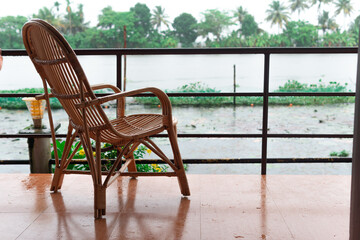 An empty cane chair overlooking the backwater in Allepey, Kerala during a rainy day in the monsoon season from a balcony near the waterfront in a luxury resort - obrazy, fototapety, plakaty