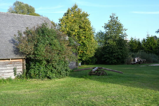 A view of an old wooden hut, shack, or stable surrounded with growing flowers, herbs, a wooden fence, and ancient farming equipment seen next to a well maintained lawn, pastureland, or meadow