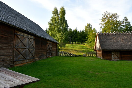 A view of an old wooden hut, shack, or stable surrounded with growing flowers, herbs, a wooden fence, and ancient farming equipment seen next to a well maintained lawn, pastureland, or meadow