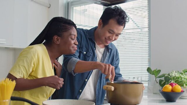 Medium shot of black woman sniffing dish while her cheerful Asian boyfriend cooking pasta in pot on stove, preparing dinner at home