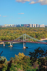 Beautiful aerial cityscape of Kyiv at sunny autumn day. Trukhaniv island with green trees. Pedestrian bridge over Dnipro river. Colorful modern skyscrapers on horizon. Travel and tourism concept