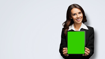 Portrait of happy smiling brunette businesswoman showing tablet pc, touchpad, with green chroma key screen, against lightly grey gray background. Cheerful beautiful business woman at office.