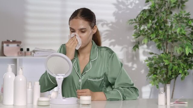 A young frustrated woman wipes off foundation or face mask with a damp napkin. The woman is upset about her problem skin. A woman in green silk pajamas sits at a dressing table in front of a mirror.
