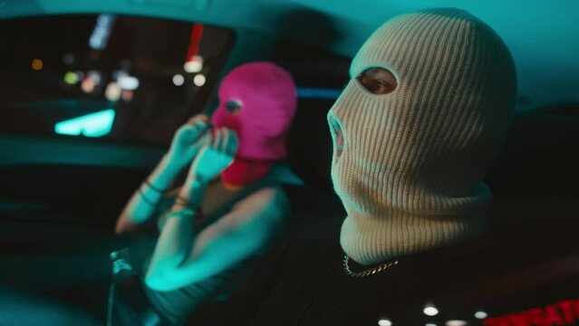 Medium side handheld shot of young Caucasian couple of robbers putting on balaclavas and taking guns in car before robbery