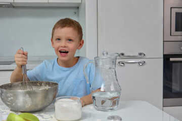 Funny Caucasian child with an open mouth cheerfully mixes ingredients for an apple pie in a deep metal bowl. Funny young cook helps in the home kitchen with the preparation of dough