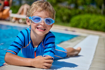 Charming little boy with curly hair in a blue bathing suit relaxes on the edge of the pool, having...