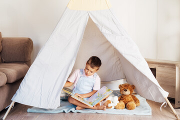 Cute little preschooler boy reading book to his toys sitting in cozy kids teepee tent in the living room at home. Child entertaining himself. Home children's activity. © okrasiuk