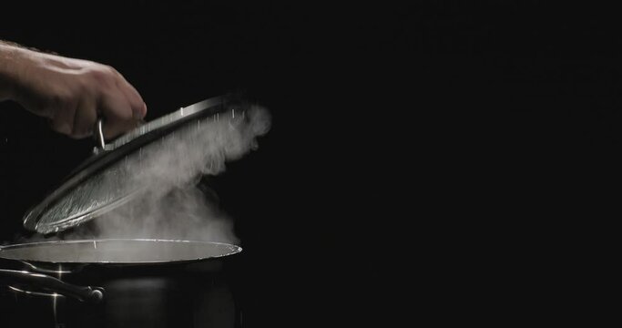 Chef opens the lid of the pan with boiling water. Steam comes out of the saucepan while cooking. Boiling water in a pan on black background. Slow motion