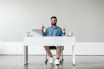 Indian businessman sitting at desk at workplace and pointing fingers up at free place above his head