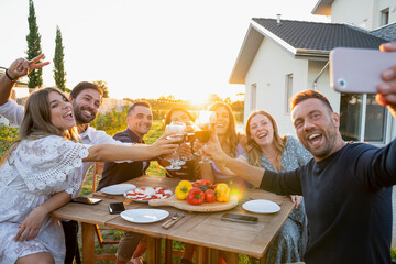 Group of friends take selfie with smartphone in garden of house toasting with glasses of red wine on a summer evening - People having fun together at party in a relaxing moment at sunset - Copy space