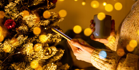 Side view of smartphone and cup in female hands in front of Christmas tree.
