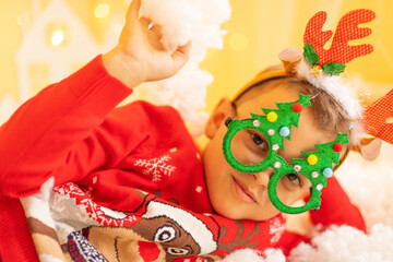 Cheerful little boy wearing warm red xmas sweater, deer horns on head and funny glasses in form of Christmas tree looks at camera and smiles on the background of New Year decorations