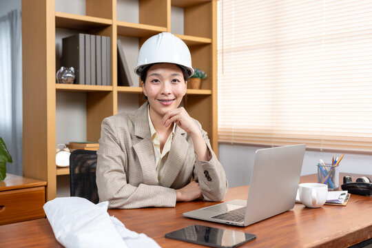 Female industrial engineer in hard hat uses laptop computer while sitting at desk Smile and be happy with the work you do.
Looking into the camera, chin resting on hand, poses confidently.
On the tabl