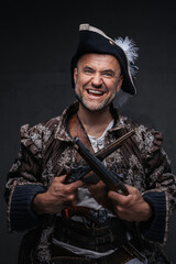 Fototapeta premium A wild-eyed pirate with a crazy smile and gray beard dressed in a vest and hat holding two muskets against a dark wall