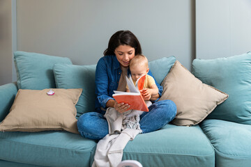 Mom reading a book with baby boy or girl at home. Early age children education, development. Mother and child spending time together read fairy tales on sofa in evening. Parent love concept - obrazy, fototapety, plakaty