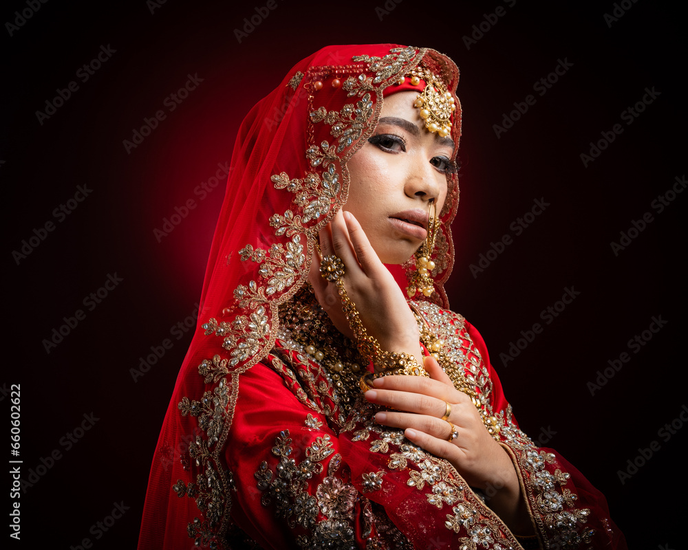 Wall mural Close up portrait of a beautiful Asian Muslim lady in a hijab wearing a gorgeous Bollywood or Indian themed red traditional wedding dress isolated on dark background