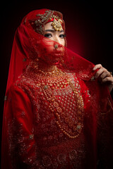 Close up portrait of a beautiful Asian Muslim lady in a hijab wearing a gorgeous Bollywood or Indian themed red traditional wedding dress isolated on dark background