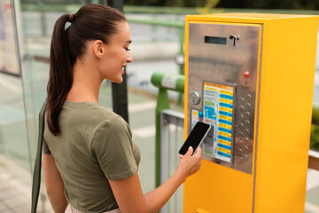 Young woman standing on bus stop paying ticket with smartphone - obrazy, fototapety, plakaty