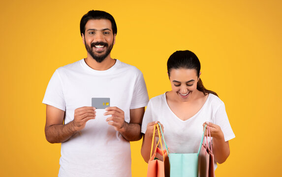 Smiling millennial arab husband show credit card, recommends sale, cusious wife look at bag with purchases