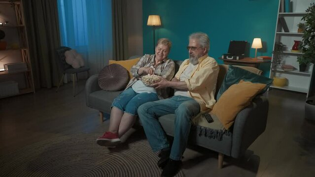 In the shot, an elderly couple are sitting on a couch watching a movie or show on TV and eating popcorn. They are laughing, giggling apparently watching a comedy. They are happy, joyful