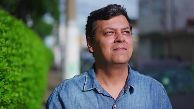 Contemplative Brazilian Man Observing Neighborhood While Walking in City Street, person feeling sunlight, face close-up in tracking shot