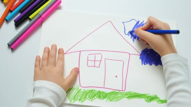 Little child drawing with felt-tip pens a house with grass and a cloud in a sketchbook.
