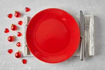 Red ceramic plate with cutlery on a white wooden tray surrounded by red Christmas balls and hearts on a light concrete table. Template for displaying New Year's food
