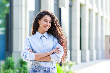 Portrait of happy and successful business woman, boss in shirt smiling and looking at camera inside office with crossed arms, Hispanic woman with curly hair outdoors