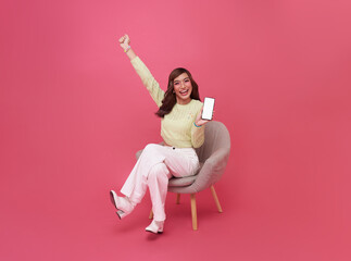 Portrait of happy Asian woman showing smartphone with white blank screen in hand sitting in armchair isolated on pink background.Gadget with empty free space for mockup, banner