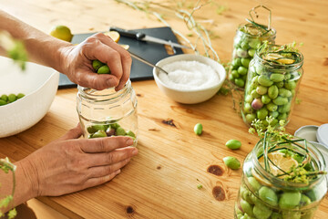 Woman preparing fermented olives in glass jar with slices of lemon, wild fennel and canning brine....