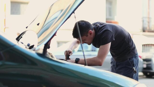 One young Caucasian man with his hands tightens a headlight bulb with a wrench in the open hood of a car during the day on a city street, close-up side view. The concept of replacing a car light bulb.