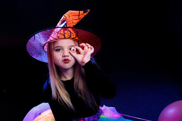 Little girl dressed as a witch with pumpkin on a dark background in the light of multi-colored...