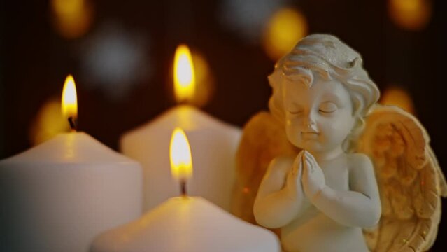 A statuette of an angel next to burning candles, against a background of garlands, taken in close-up. Merry Christmas. A request in prayer.