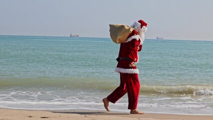 Santa Claus with a bag of gifts on his shoulder walks along the sandy beach along the surf line on a hot summer day. Christmas or New Year holidays in hot countries. Santa's summer vacation.