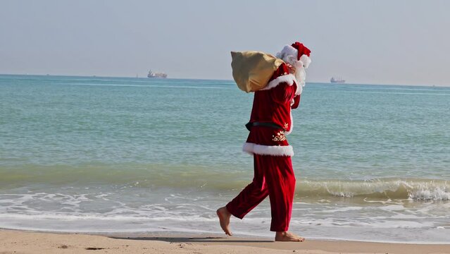 Santa Claus with a bag of gifts on his shoulder walks along the sandy beach along the surf line on a hot summer day. Christmas or New Year holidays in hot countries. Santa's summer vacation.