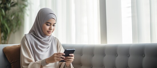 Beautiful Muslim woman playing with cellphone sitting on sofa chair - obrazy, fototapety, plakaty