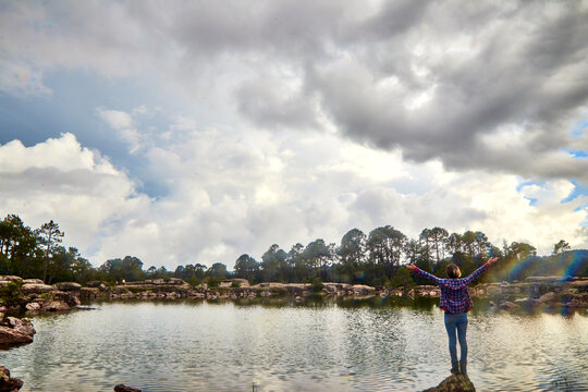 woman standing up with arms up in front of a lake in rainny forest, cloud sky in mountains at summer 