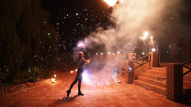 Female artist spins burning staff with sparks at fire performance show at night. Young woman performing amazing fire art open air on the festival. Fire dancer working with pyrotechnics on the street.