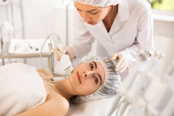Young woman cosmetologist performs facial exfoliating procedure with machine to young female client